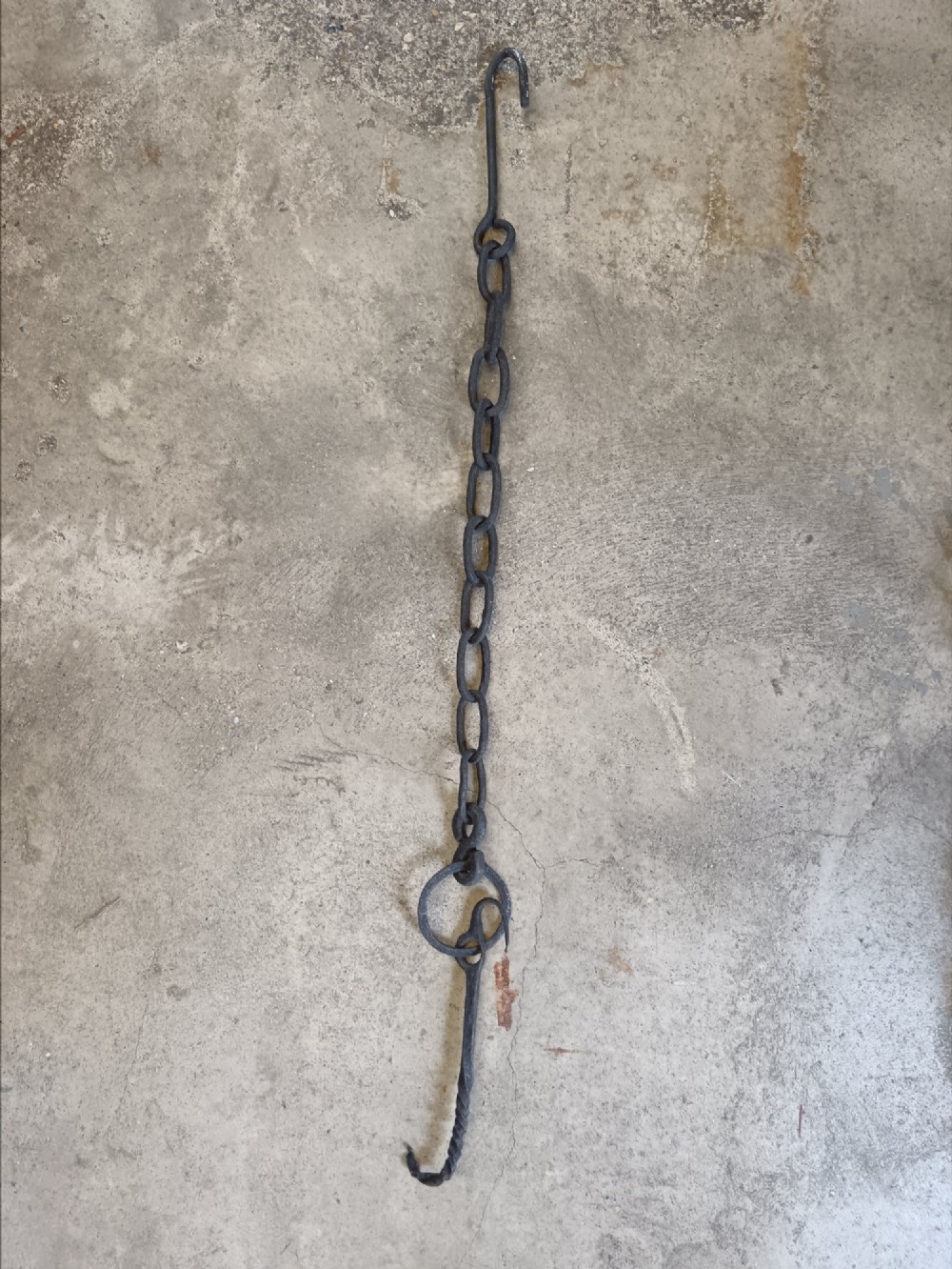 A collection of six hand-forged dark wrought iron adjustable chimney trammel hooks and pot hangers laid flat on a concrete background. View VIII
