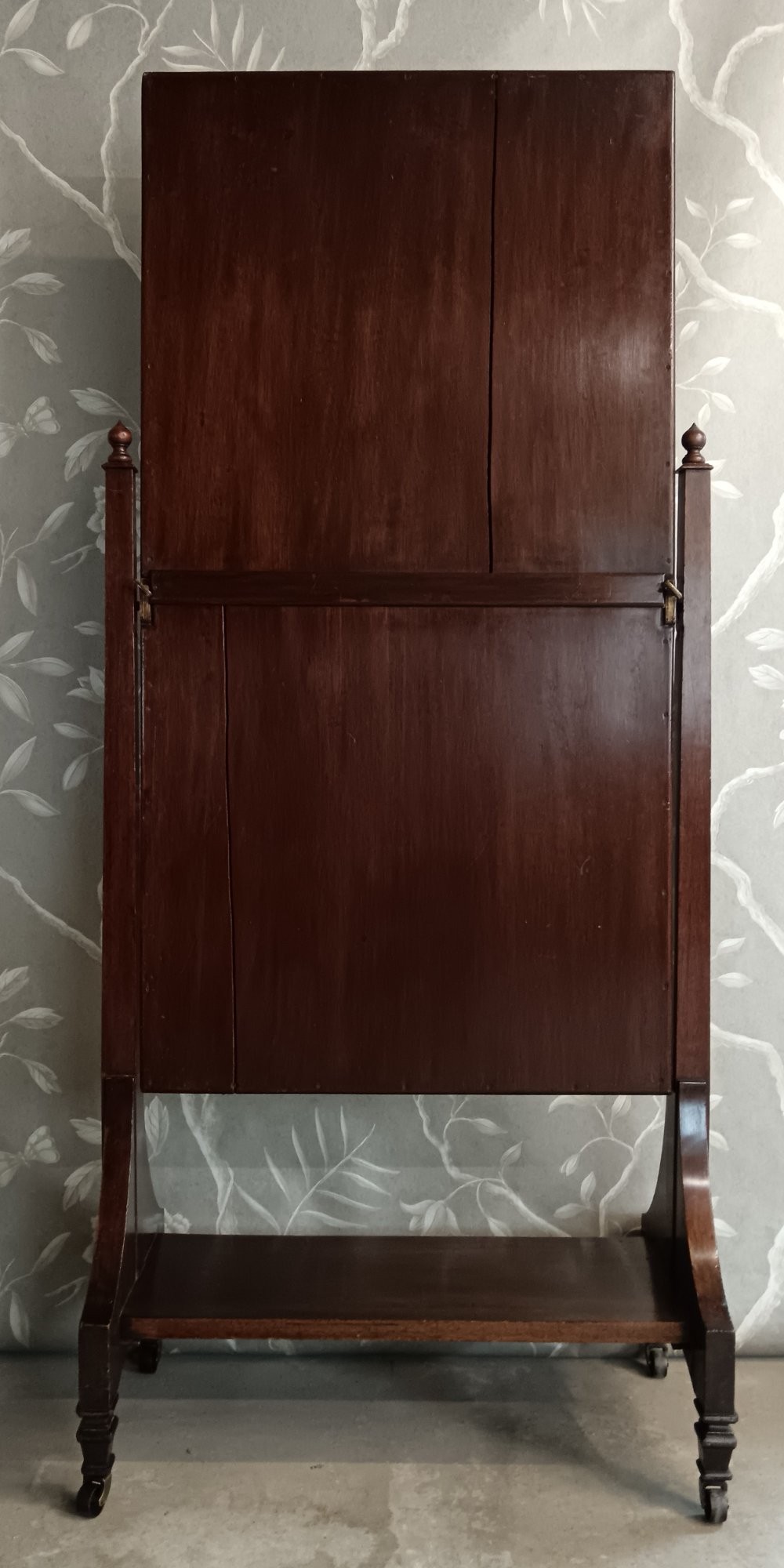 A late Victorian mahogany cheval mirror featuring a rectangular full-length pivoting frame set between turned finial posts on a shelf base with brass castors. View II