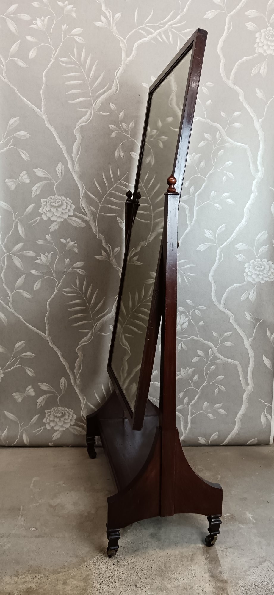 A late Victorian mahogany cheval mirror featuring a rectangular full-length pivoting frame set between turned finial posts on a shelf base with brass castors. View V