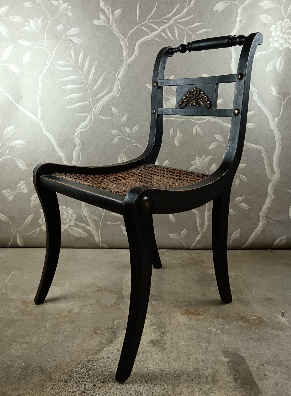 An ebonised Regency side chair featuring curved sabre legs, a woven cane seat, turned top rail, and a carved back splat with brass detail.