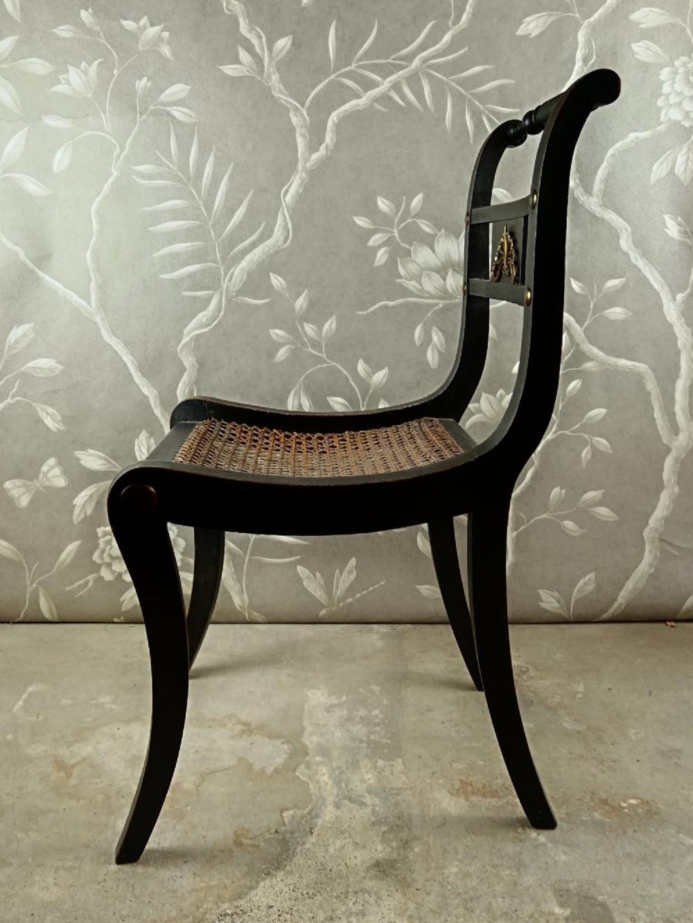 An ebonised Regency side chair featuring curved sabre legs, a woven cane seat, turned top rail, and a carved back splat with brass detail. View II