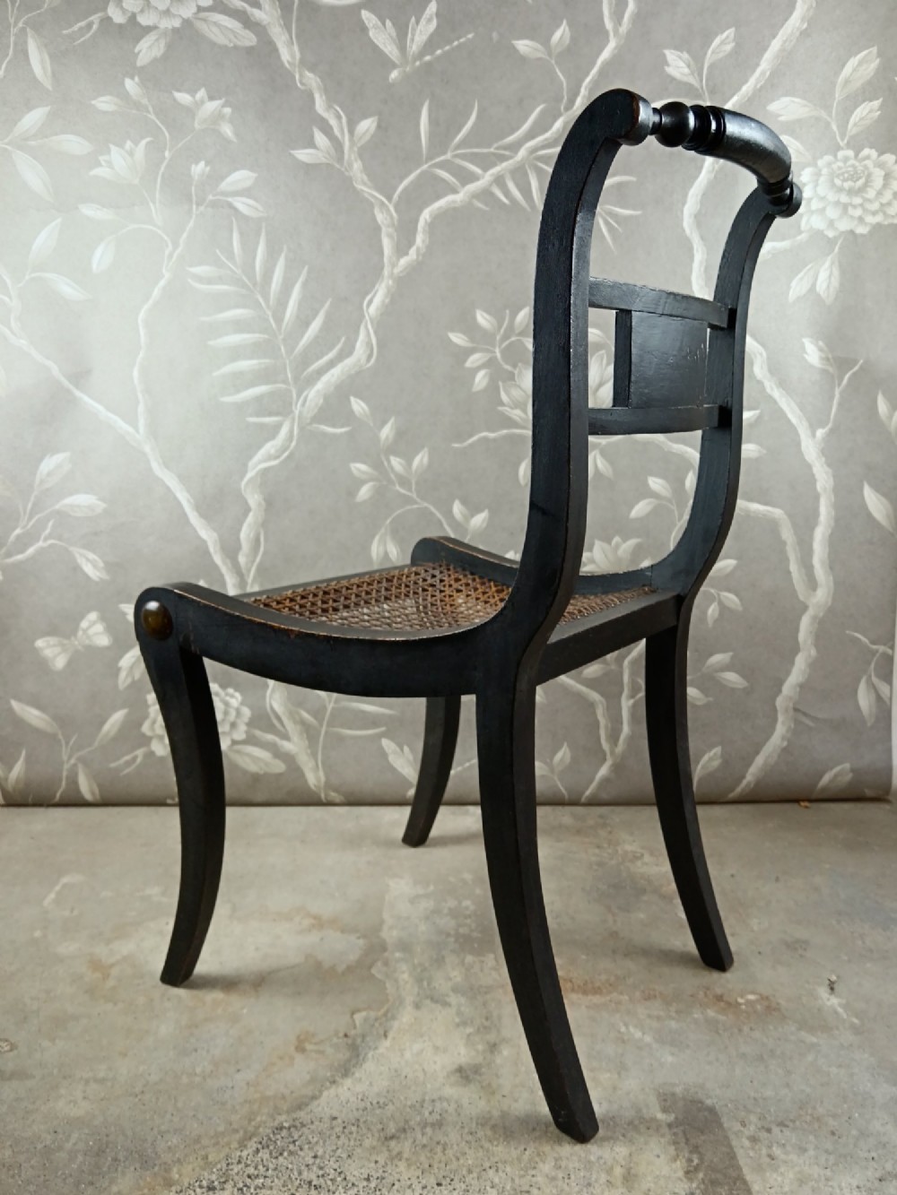 An ebonised Regency side chair featuring curved sabre legs, a woven cane seat, turned top rail, and a carved back splat with brass detail. View III
