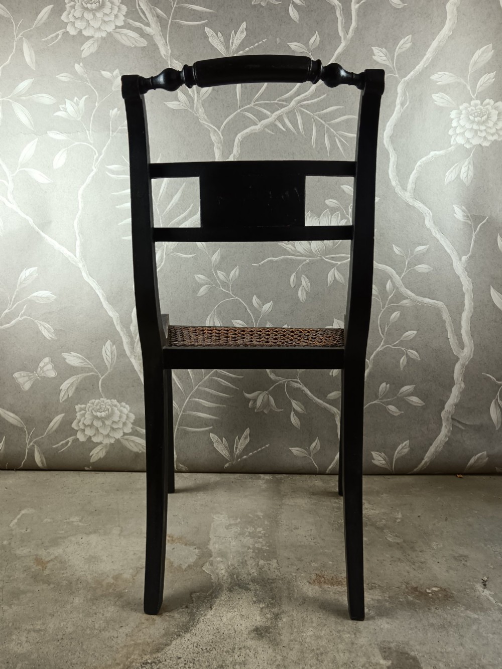 An ebonised Regency side chair featuring curved sabre legs, a woven cane seat, turned top rail, and a carved back splat with brass detail. View IV