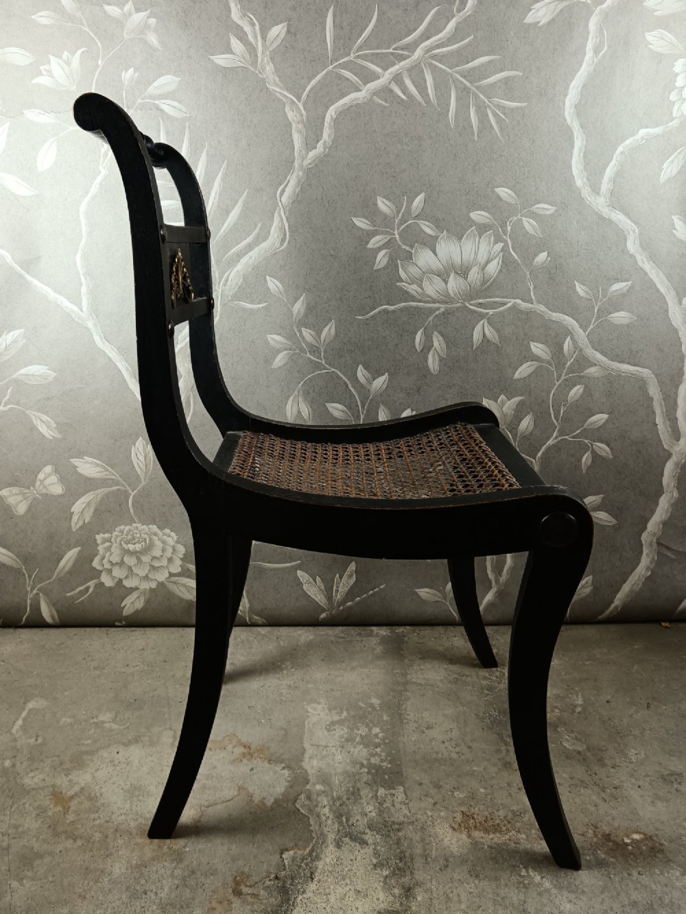 An ebonised Regency side chair featuring curved sabre legs, a woven cane seat, turned top rail, and a carved back splat with brass detail. View V