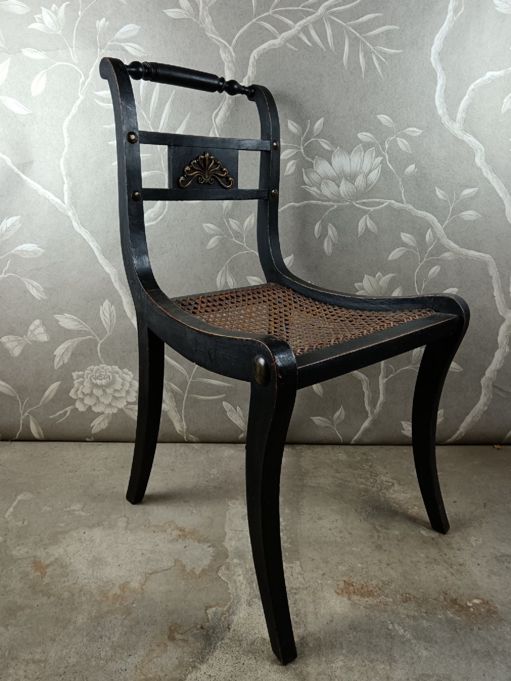 An ebonised Regency side chair featuring curved sabre legs, a woven cane seat, turned top rail, and a carved back splat with brass detail. View VI