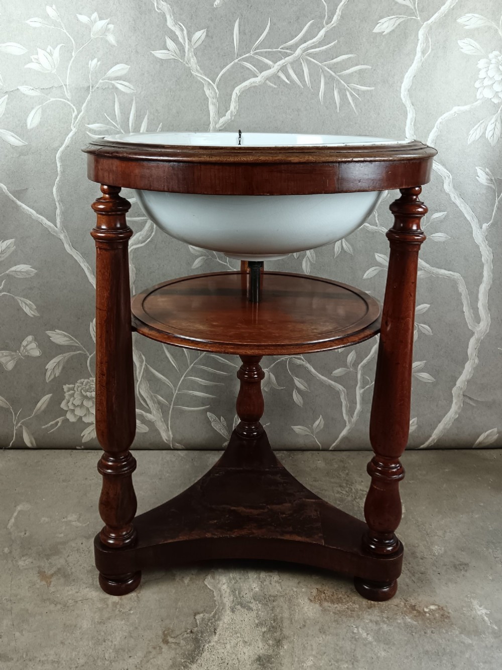 A 19th-century circular mahogany campaign washstand featuring three turned legs, a circular tier, a triangular platform base, and an inset white ceramic wash basin.