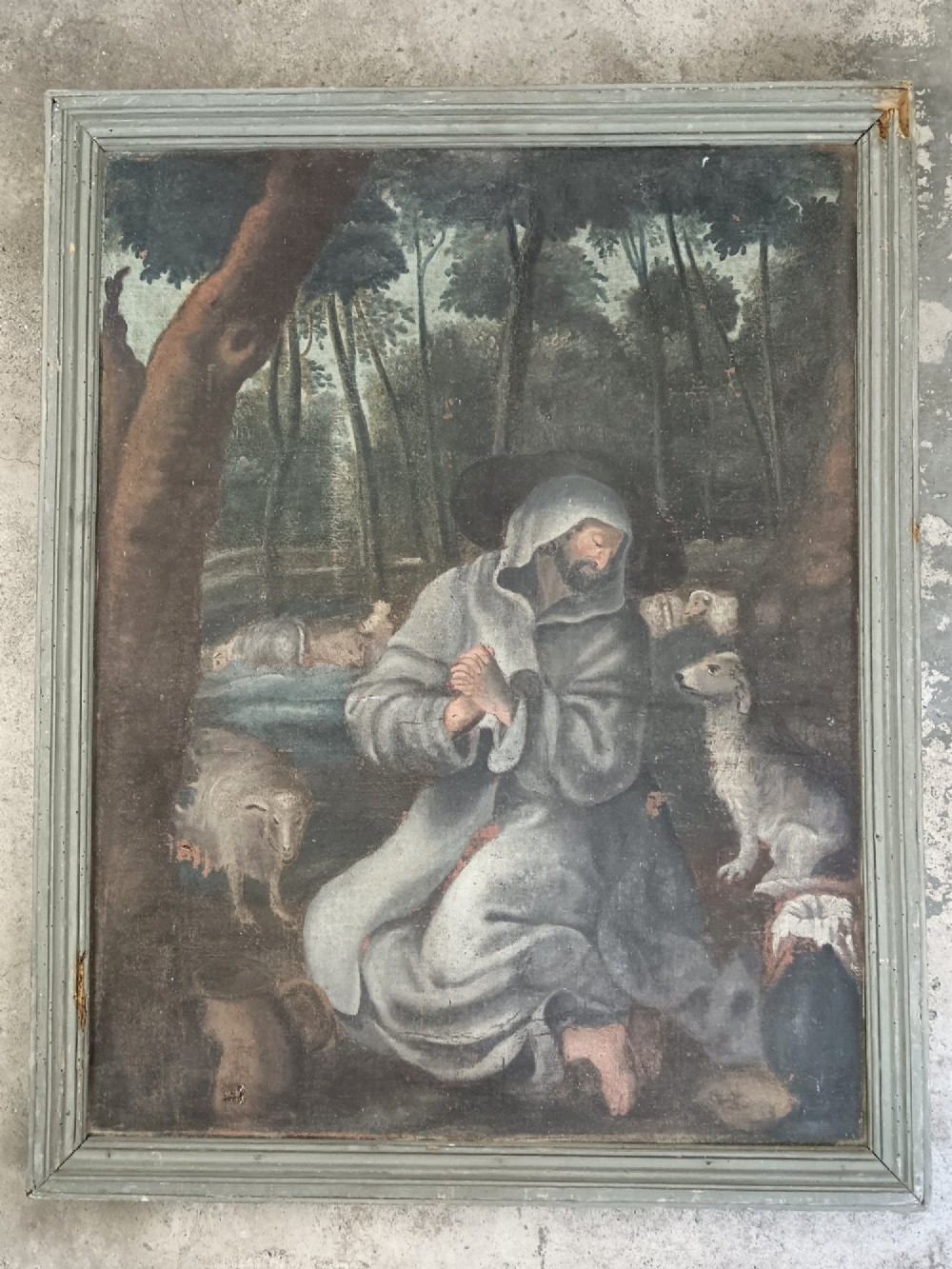 An antique naive oil painting depicting Saint Francis kneeling in prayer amongst sheep in a wooded landscape within a grey painted frame.