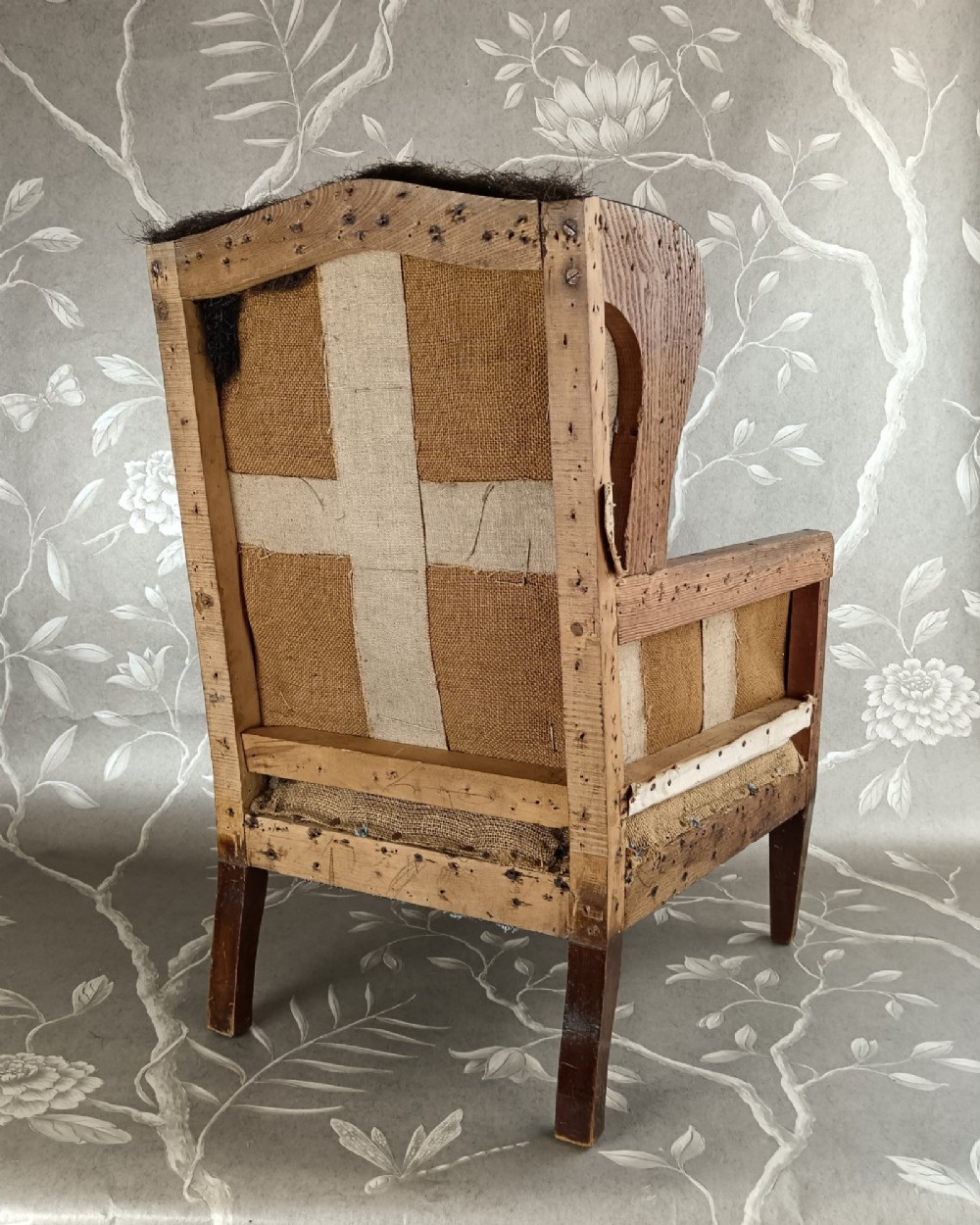 An early 20th-century childs wingback armchair featuring an exposed timber frame, tapered legs, and visible hessian upholstery layer. View V