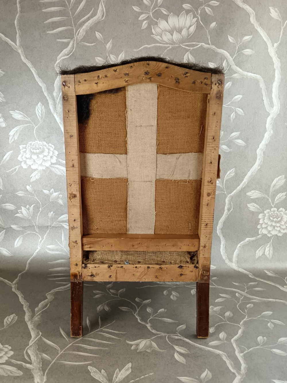 An early 20th-century childs wingback armchair featuring an exposed timber frame, tapered legs, and visible hessian upholstery layer. View VI