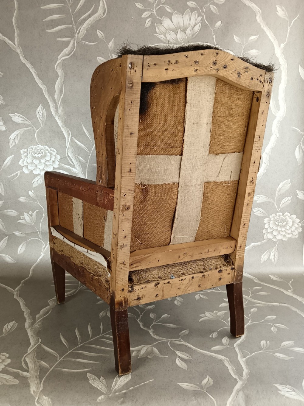 An early 20th-century childs wingback armchair featuring an exposed timber frame, tapered legs, and visible hessian upholstery layer. View VII