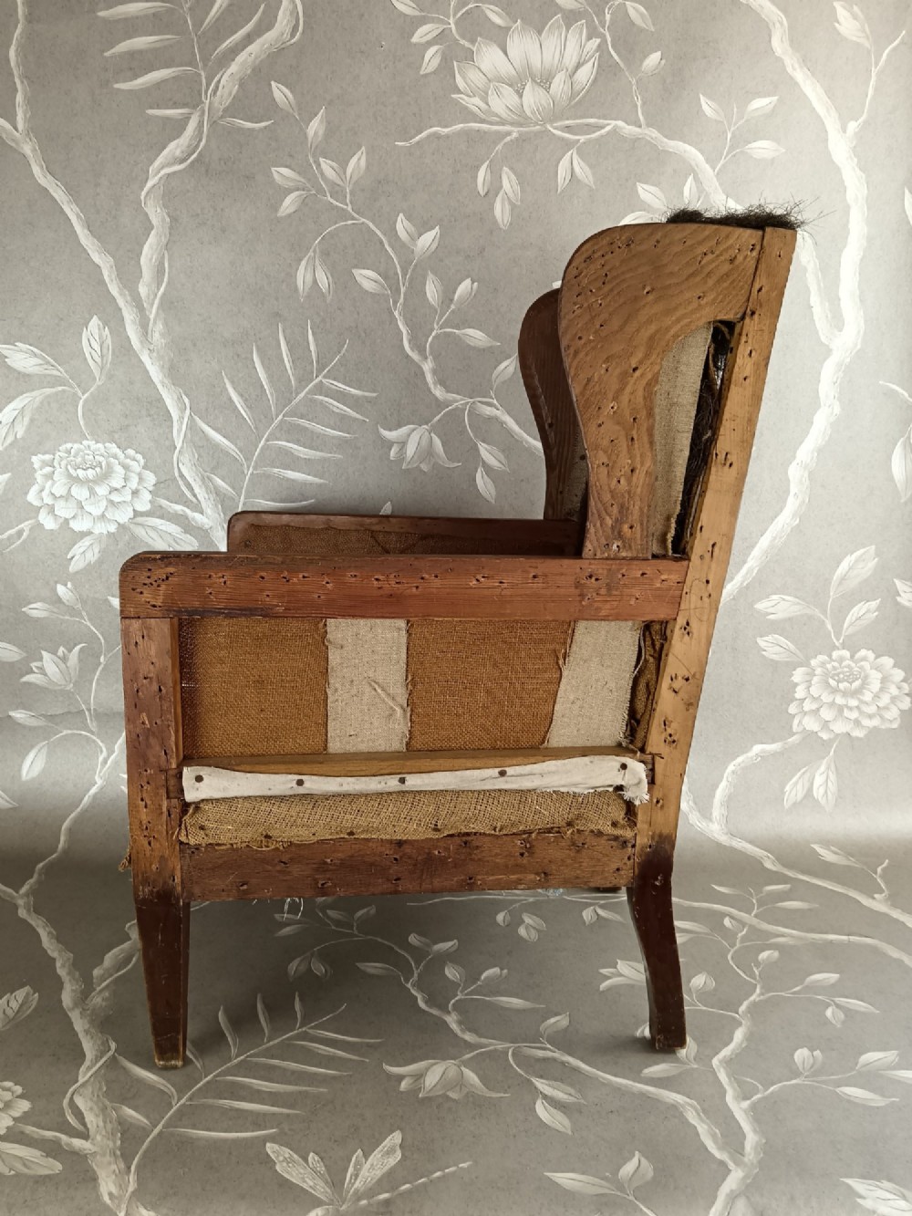 An early 20th-century childs wingback armchair featuring an exposed timber frame, tapered legs, and visible hessian upholstery layer. View VIII