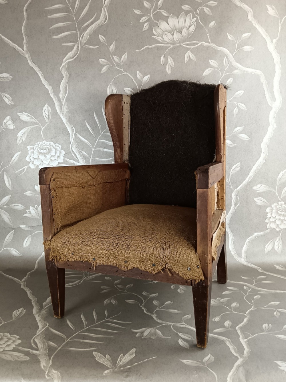 An early 20th-century childs wingback armchair featuring an exposed timber frame, tapered legs, and visible hessian upholstery layer. View IX