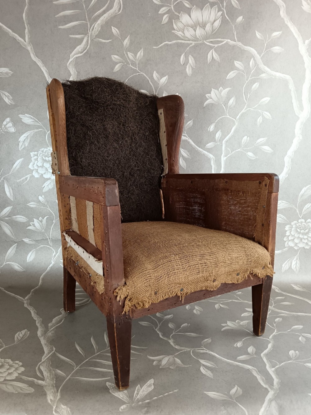 An early 20th-century childs wingback armchair featuring an exposed timber frame, tapered legs, and visible hessian upholstery layer. View X