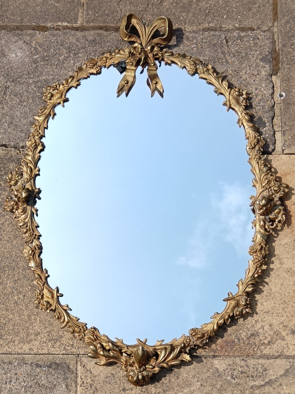 An oval wall mirror framed in detailed gilt bronze with floral scrollwork and a prominent ribbon bow crest at the top. View II