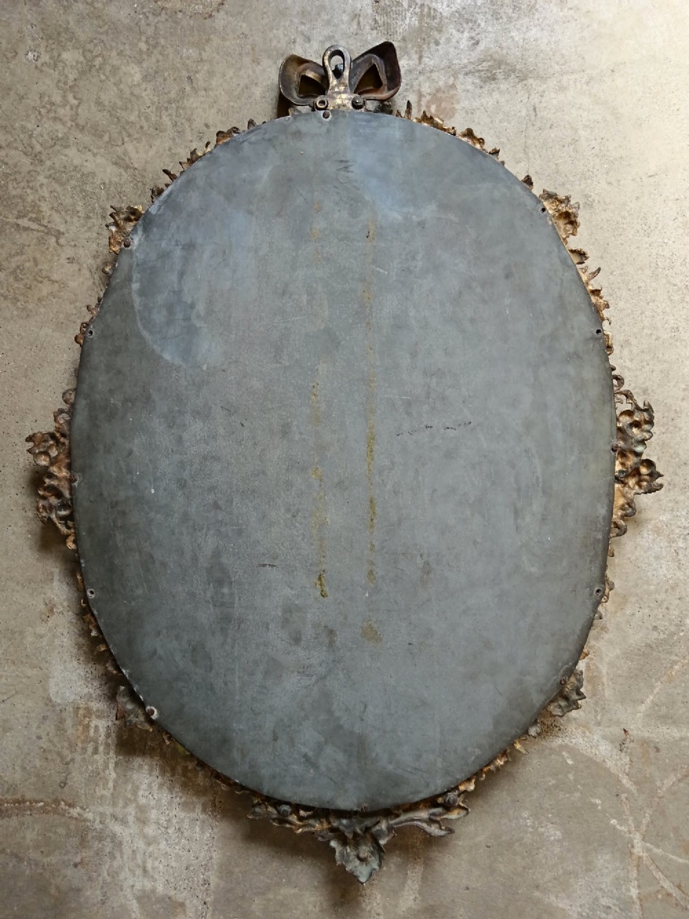An oval wall mirror framed in detailed gilt bronze with floral scrollwork and a prominent ribbon bow crest at the top. View VII