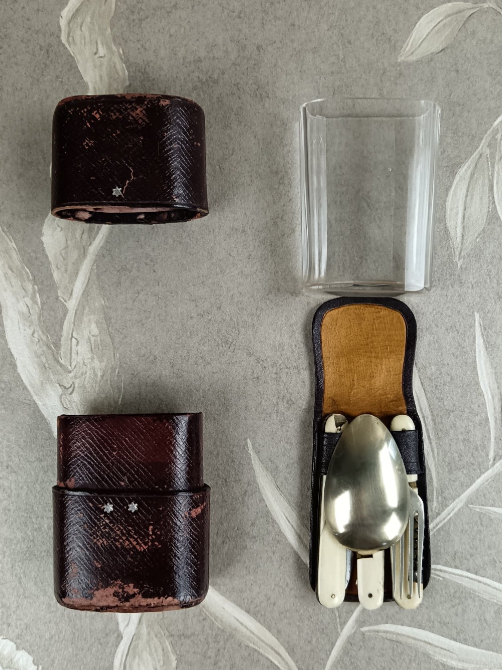 A Victorian folding campaign cutlery set featuring silver-plated utensils with light-coloured handles, a clear glass beaker, and a fitted dark red morocco leather carrying case. View IV