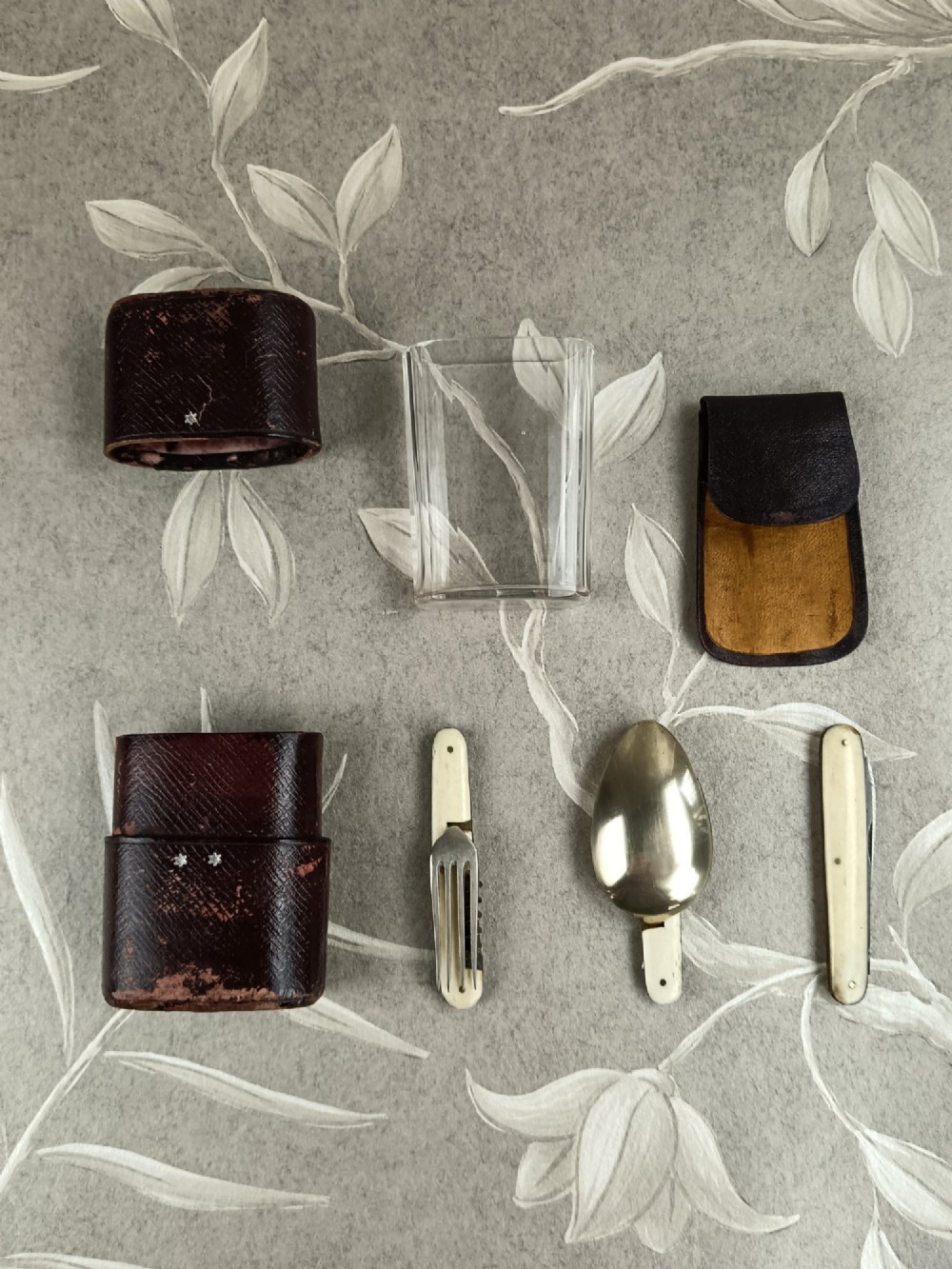 A Victorian folding campaign cutlery set featuring silver-plated utensils with light-coloured handles, a clear glass beaker, and a fitted dark red morocco leather carrying case. View V