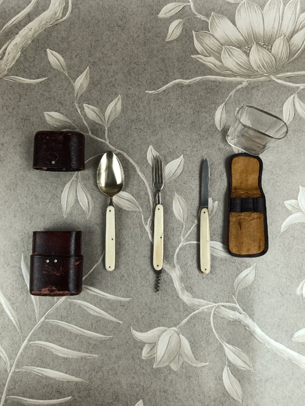 A Victorian folding campaign cutlery set featuring silver-plated utensils with light-coloured handles, a clear glass beaker, and a fitted dark red morocco leather carrying case. View VI