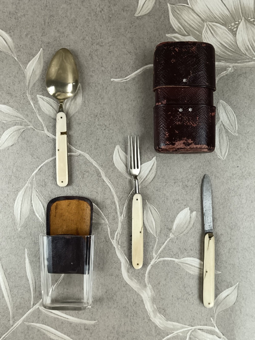 A Victorian folding campaign cutlery set featuring silver-plated utensils with light-coloured handles, a clear glass beaker, and a fitted dark red morocco leather carrying case. View VII