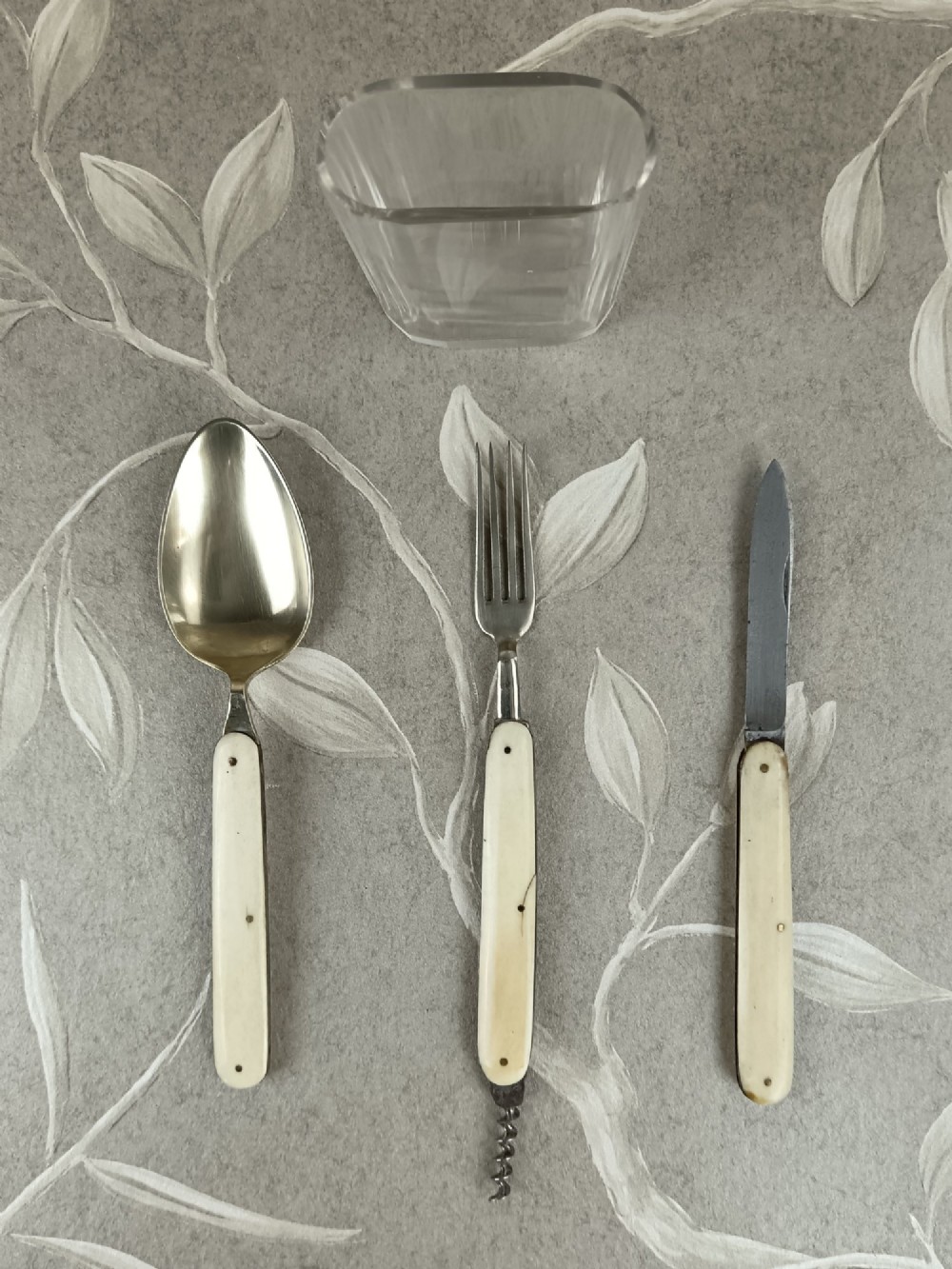 A Victorian folding campaign cutlery set featuring silver-plated utensils with light-coloured handles, a clear glass beaker, and a fitted dark red morocco leather carrying case. View IX