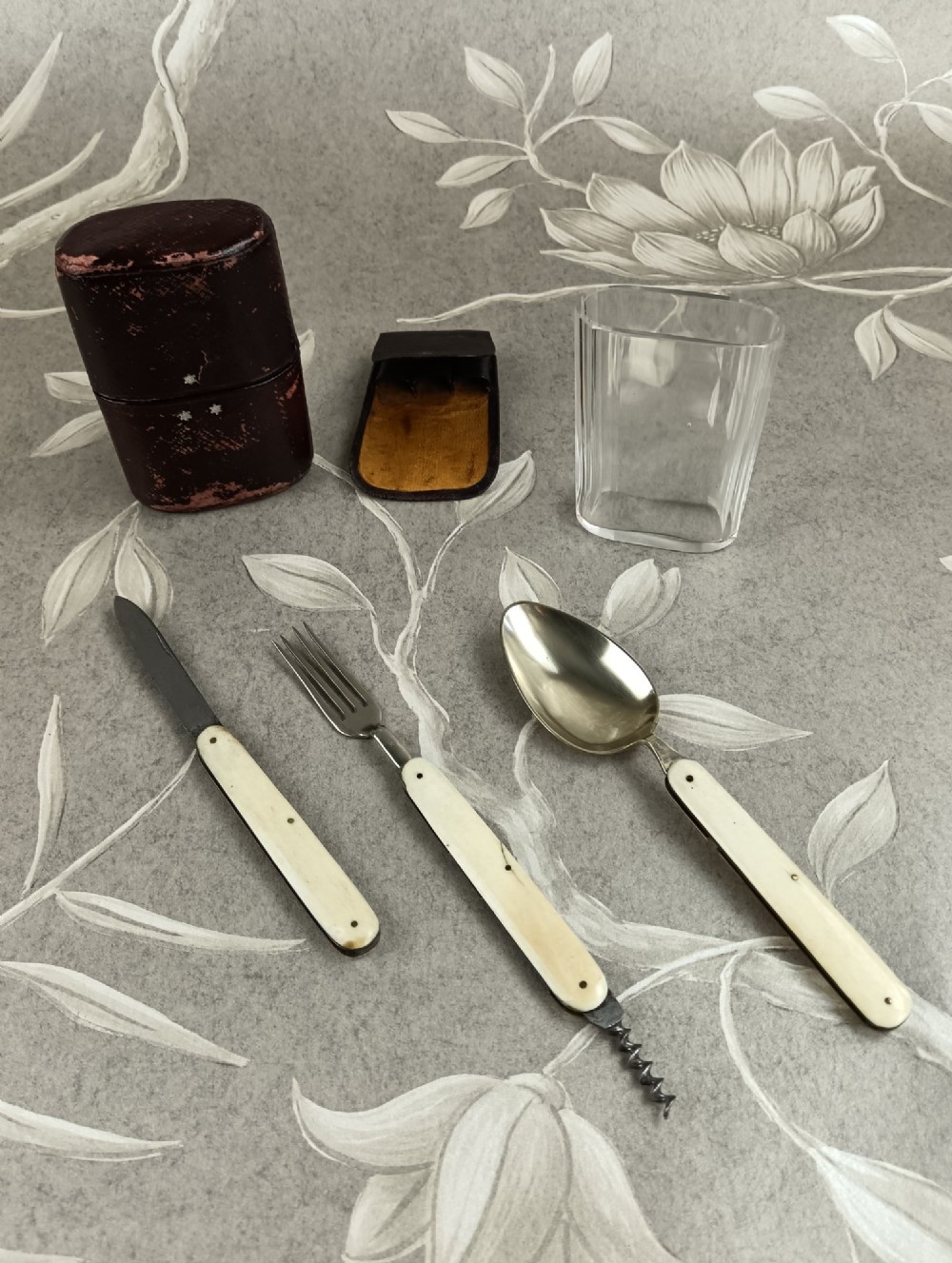 A Victorian folding campaign cutlery set featuring silver-plated utensils with light-coloured handles, a clear glass beaker, and a fitted dark red morocco leather carrying case. View X