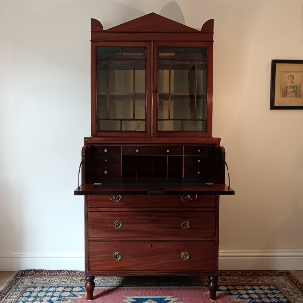 A Regency mahogany secretaire cabinet featuring a triangular pediment top, glazed upper doors, a fall-front desk section, and three drawers below on turned feet.