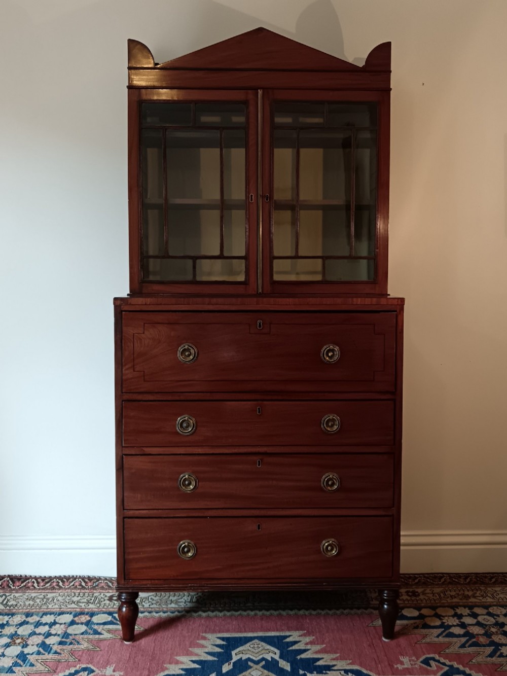 A Regency mahogany secretaire cabinet featuring a triangular pediment top, glazed upper doors, a fall-front desk section, and three drawers below on turned feet. View II