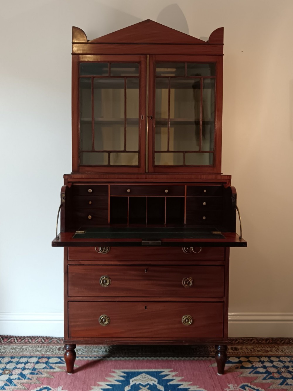 A Regency mahogany secretaire cabinet featuring a triangular pediment top, glazed upper doors, a fall-front desk section, and three drawers below on turned feet. View III