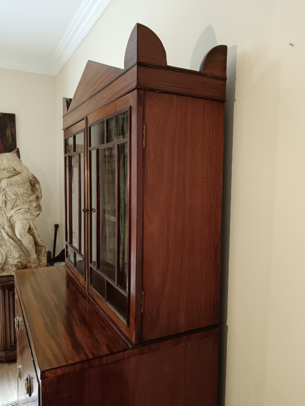 A Regency mahogany secretaire cabinet featuring a triangular pediment top, glazed upper doors, a fall-front desk section, and three drawers below on turned feet. View IX