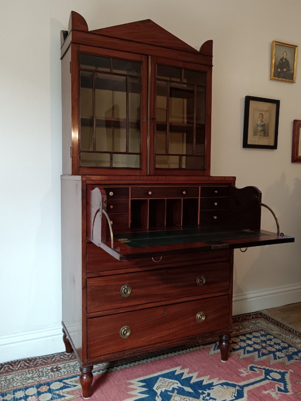 A Regency mahogany secretaire cabinet featuring a triangular pediment top, glazed upper doors, a fall-front desk section, and three drawers below on turned feet. View XI