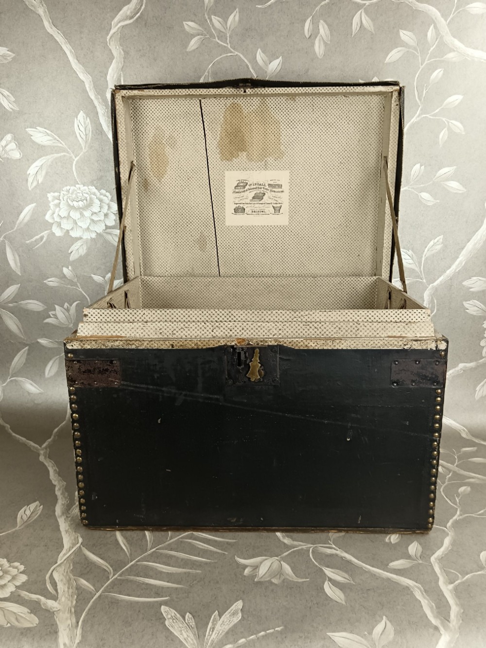 A rectangular black leather travelling trunk featuring brass stud border detail, iron corner reinforcement brackets, and a brass central lock escutcheon. View V