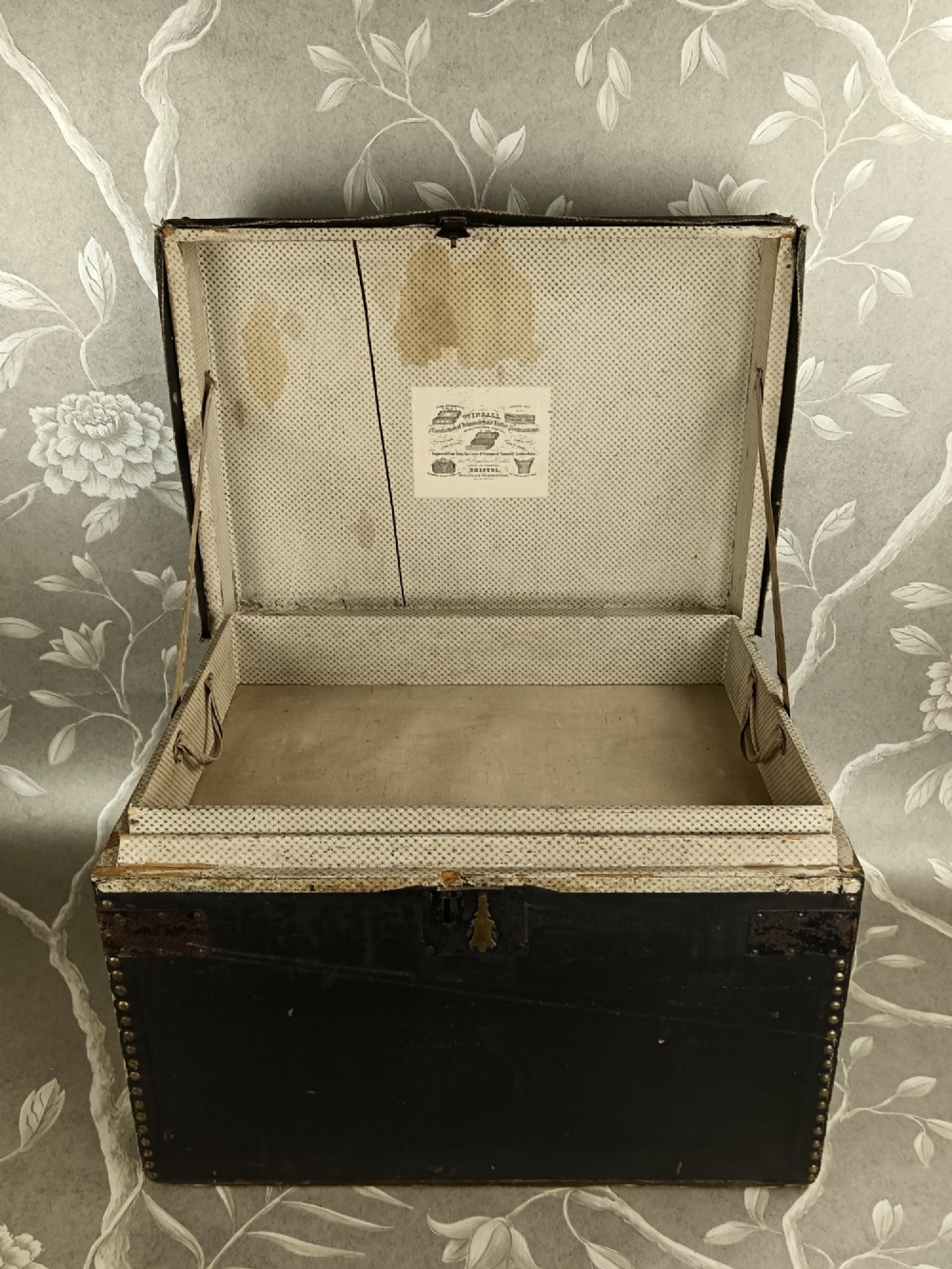 A rectangular black leather travelling trunk featuring brass stud border detail, iron corner reinforcement brackets, and a brass central lock escutcheon. View VI