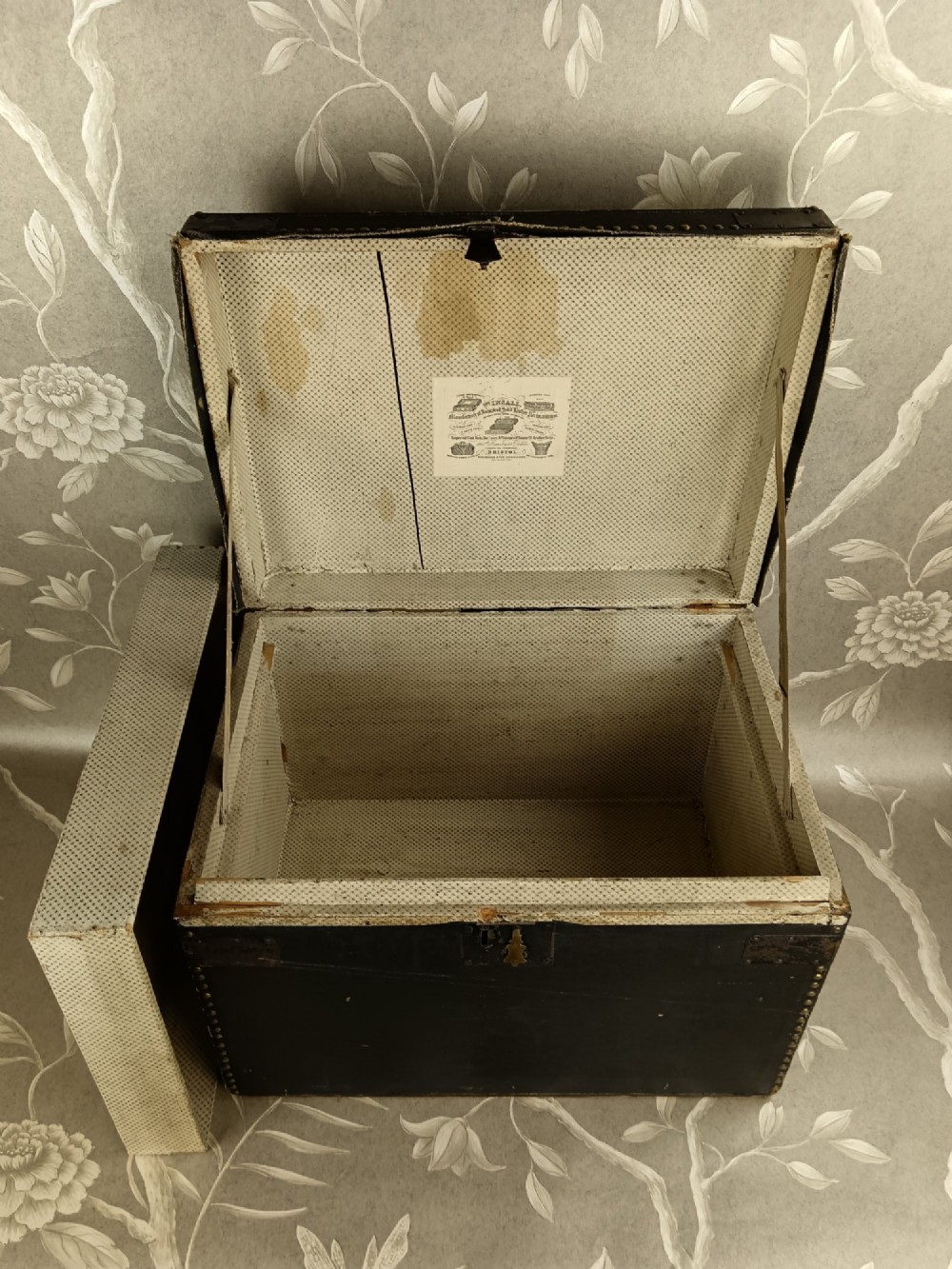 A rectangular black leather travelling trunk featuring brass stud border detail, iron corner reinforcement brackets, and a brass central lock escutcheon. View VII