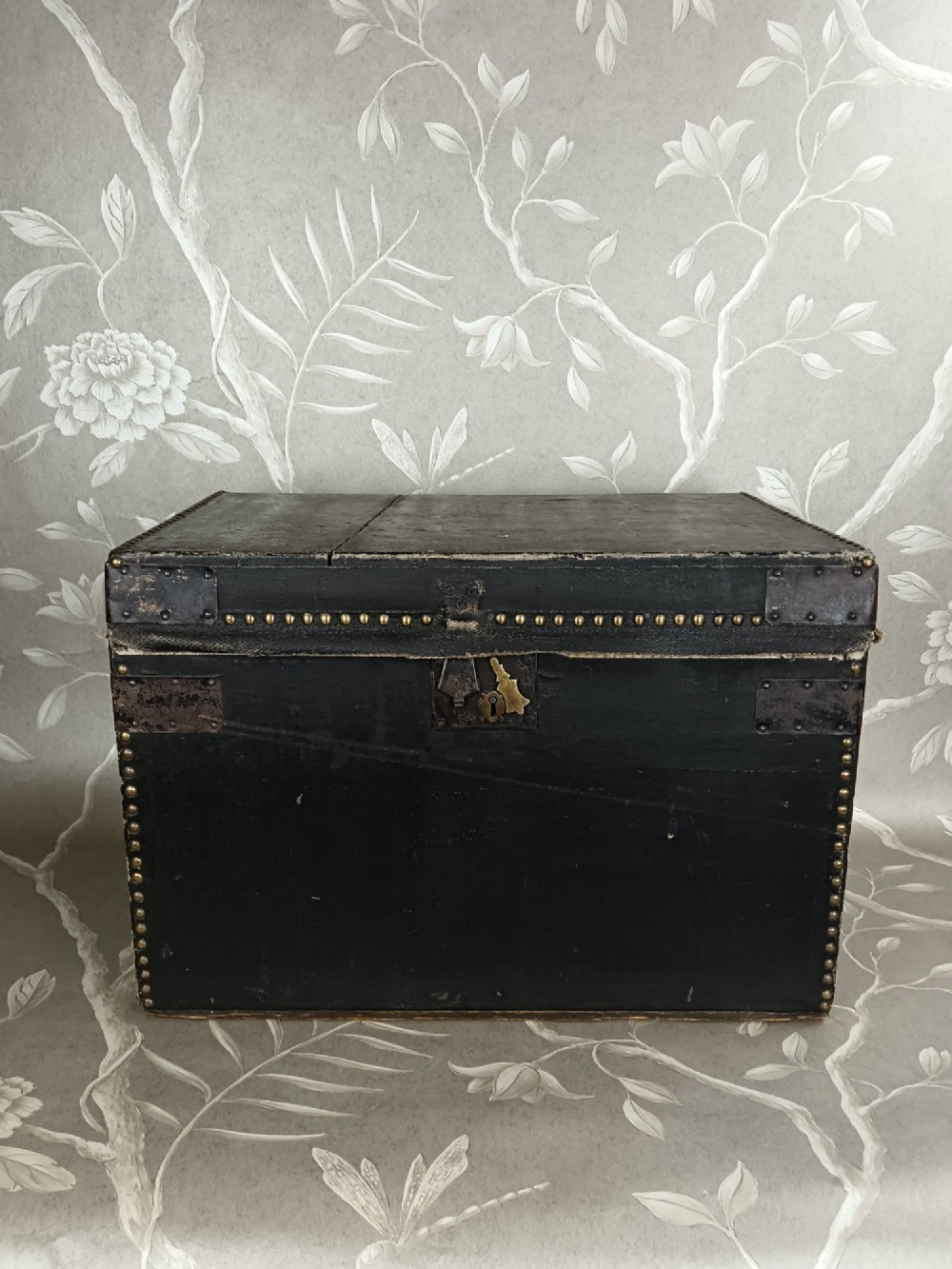 A rectangular black leather travelling trunk featuring brass stud border detail, iron corner reinforcement brackets, and a brass central lock escutcheon. View X