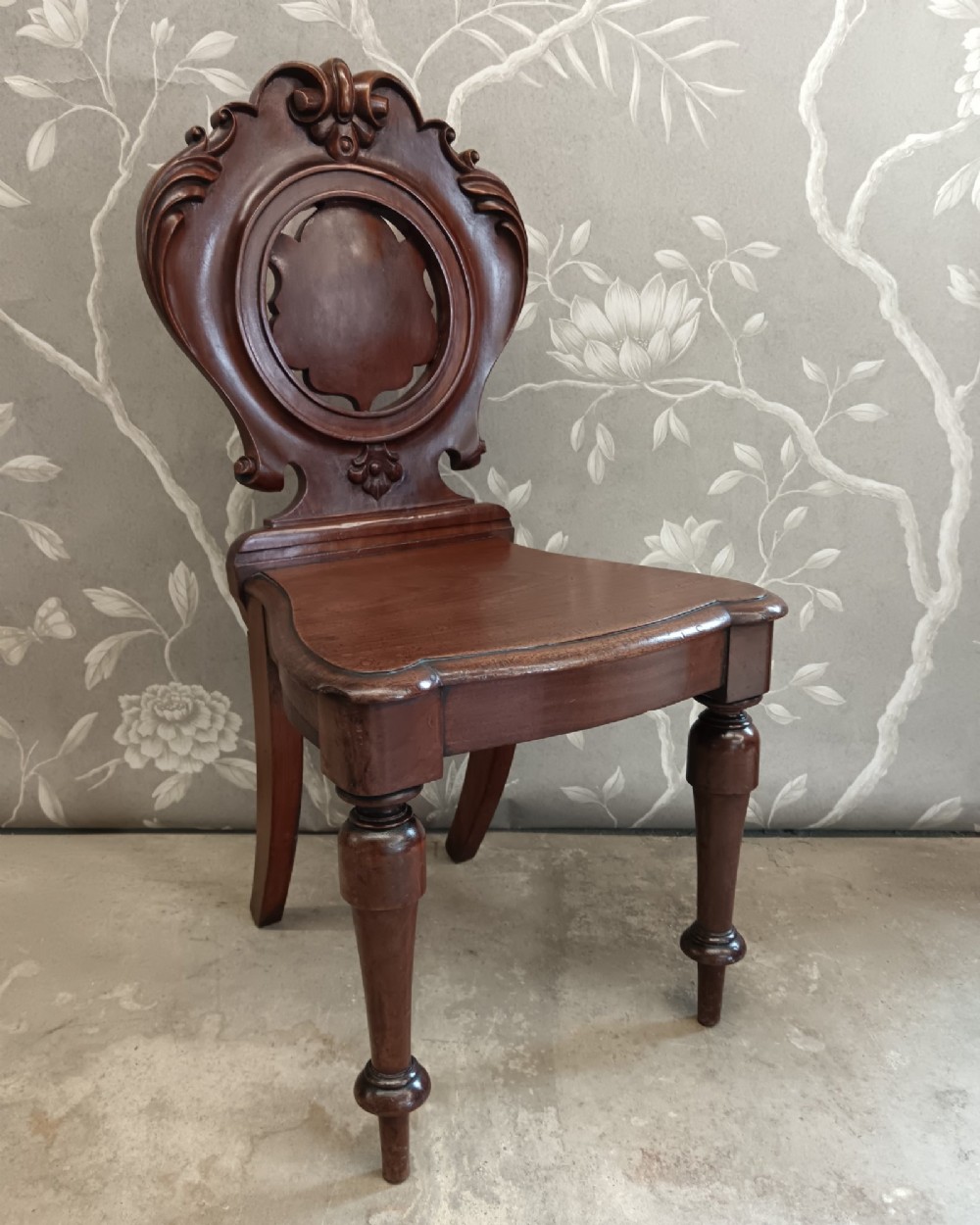 A Victorian mahogany hall chair featuring a carved shield backrest with scroll detailing, a solid wooden seat, and turned front legs. View VII