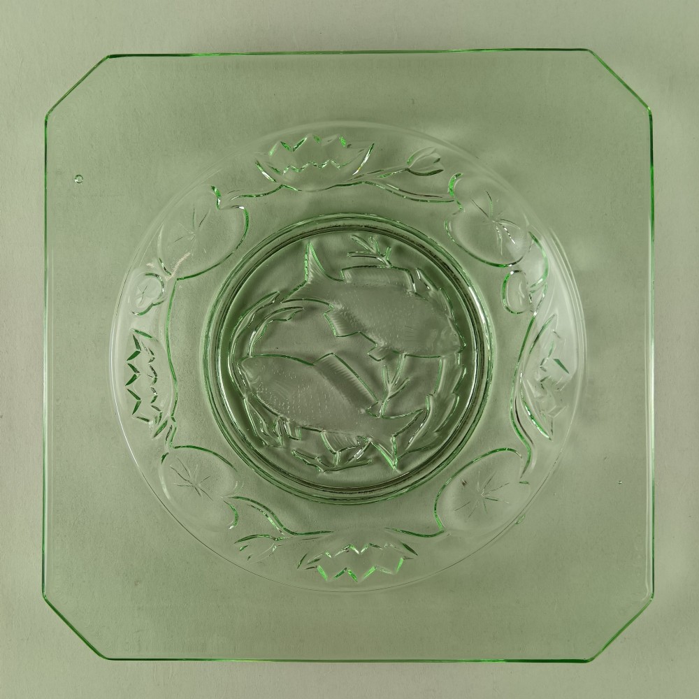 A square pale green Art Deco pressed glass plate featuring a central circular design of two swimming fish surrounded by embossed water lilies.