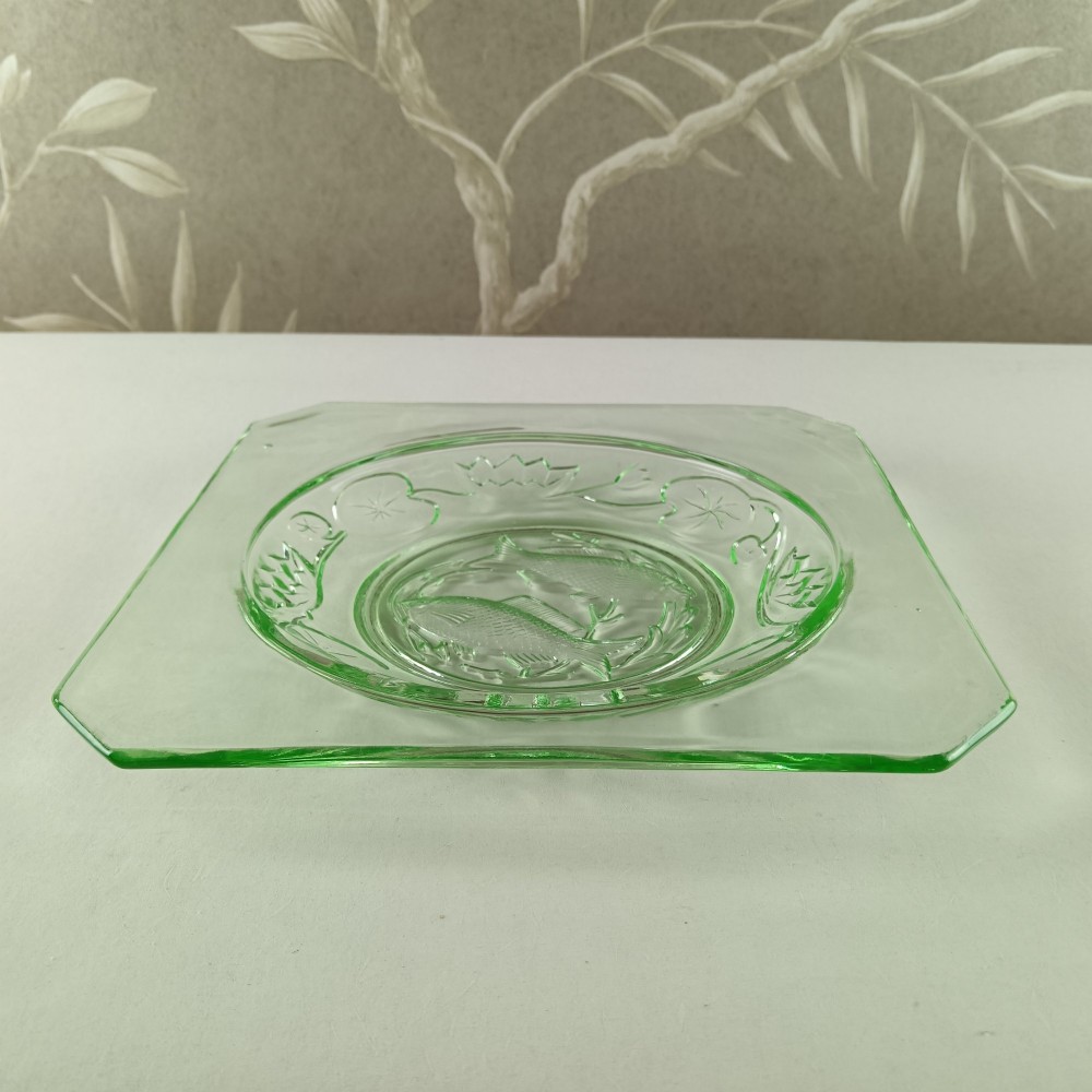 A square pale green Art Deco pressed glass plate featuring a central circular design of two swimming fish surrounded by embossed water lilies. View III