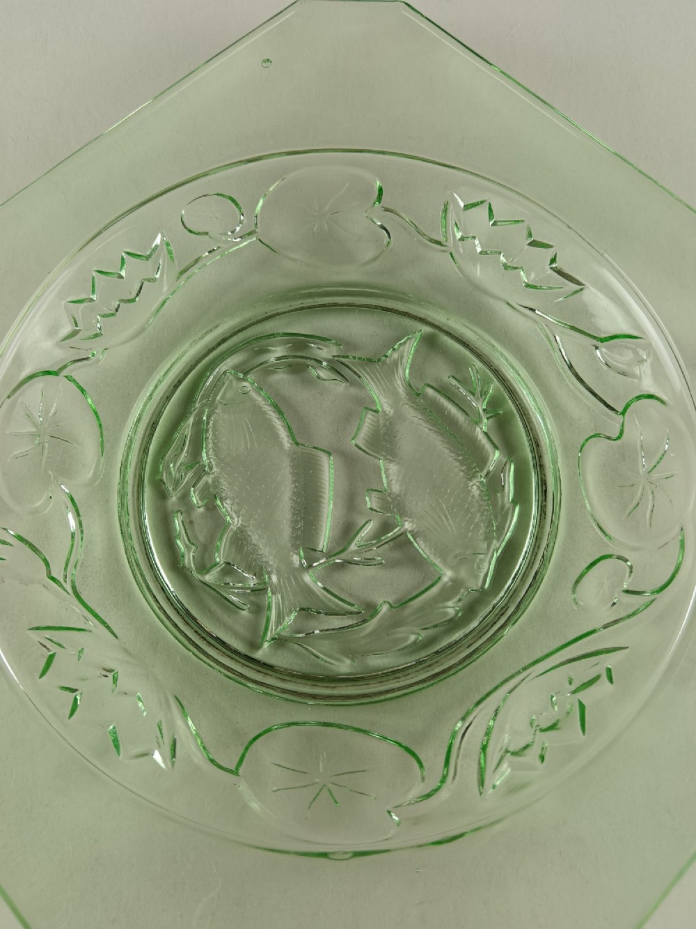 A square pale green Art Deco pressed glass plate featuring a central circular design of two swimming fish surrounded by embossed water lilies. View VII