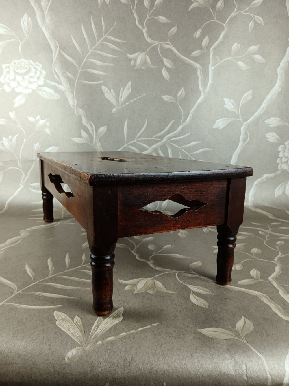 A low 19th-century dark brown wooden footstool with turned legs, side cutouts, and a central carry handle slot on the top surface. View III