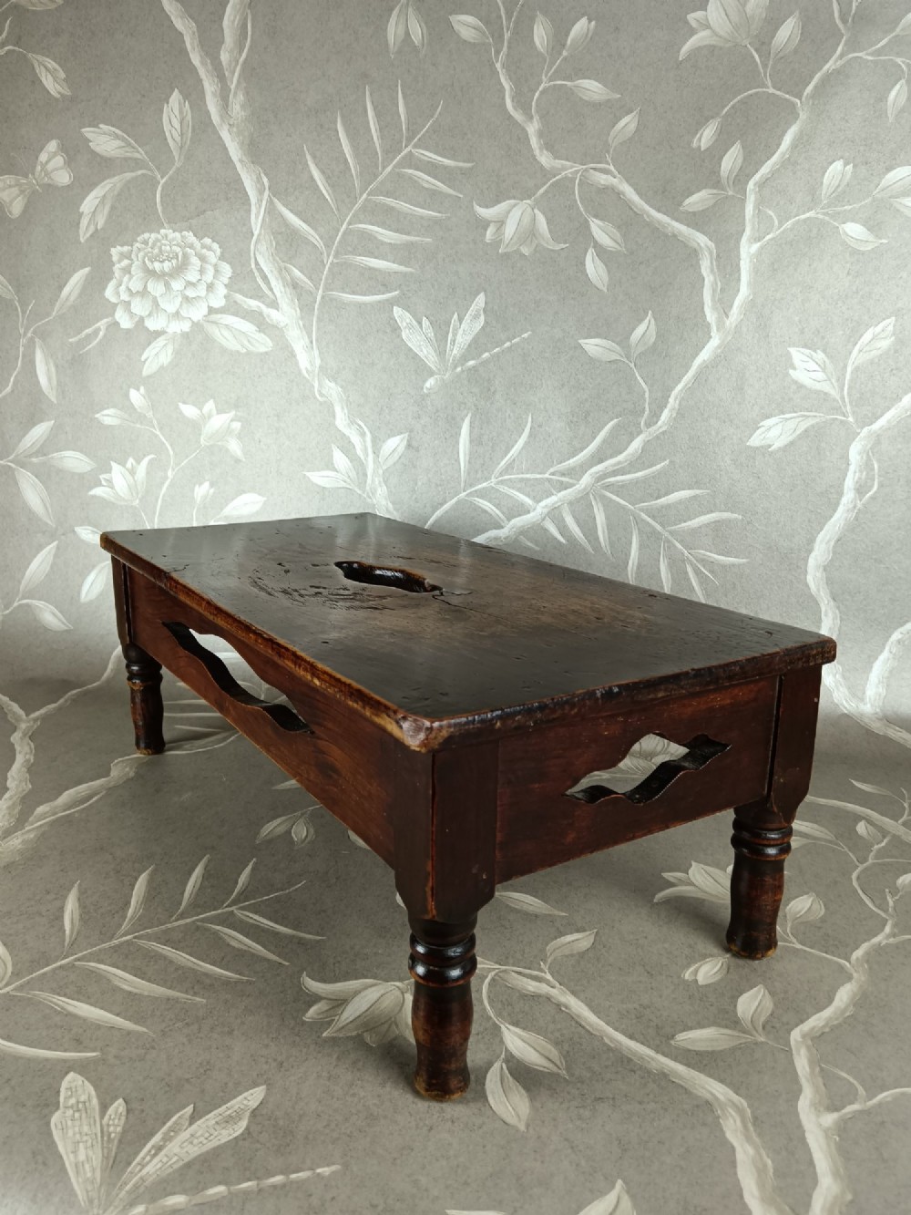 A low 19th-century dark brown wooden footstool with turned legs, side cutouts, and a central carry handle slot on the top surface. View V