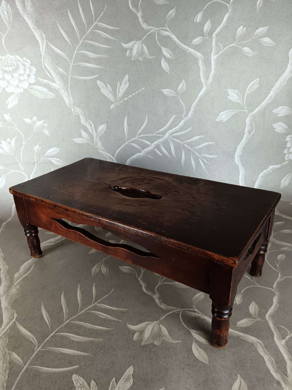 A low 19th-century dark brown wooden footstool with turned legs, side cutouts, and a central carry handle slot on the top surface. View VI