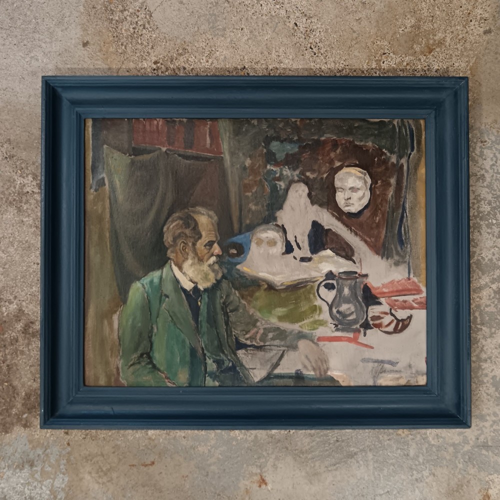 A mid-twentieth-century modernist oil painting on card depicting a bearded man in a green jacket seated in an artist studio, presented in a dark blue wooden frame.