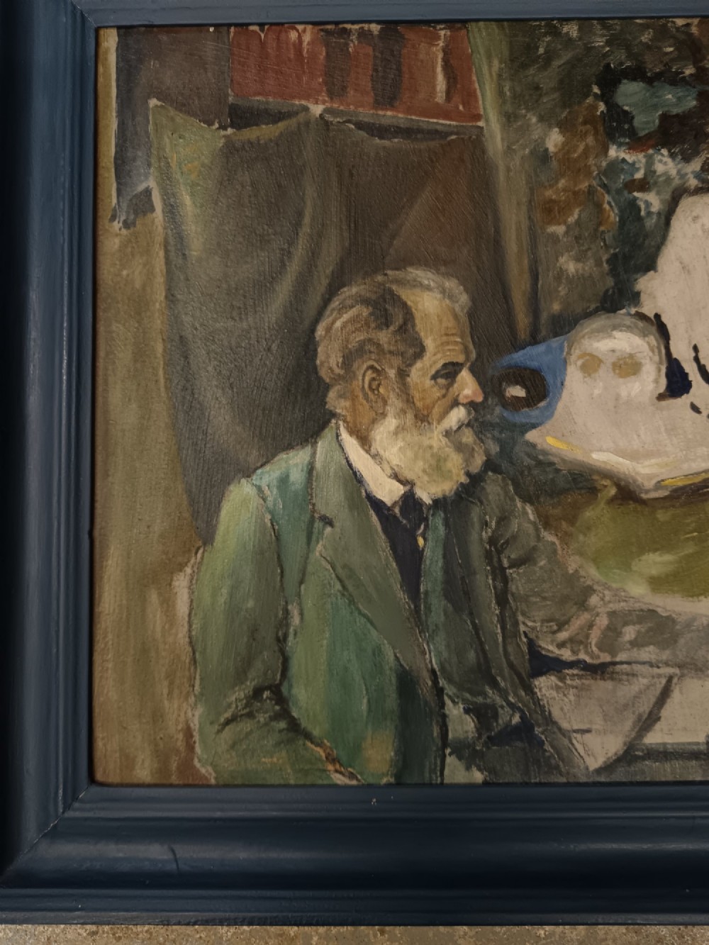 A mid-twentieth-century modernist oil painting on card depicting a bearded man in a green jacket seated in an artist studio, presented in a dark blue wooden frame. View III
