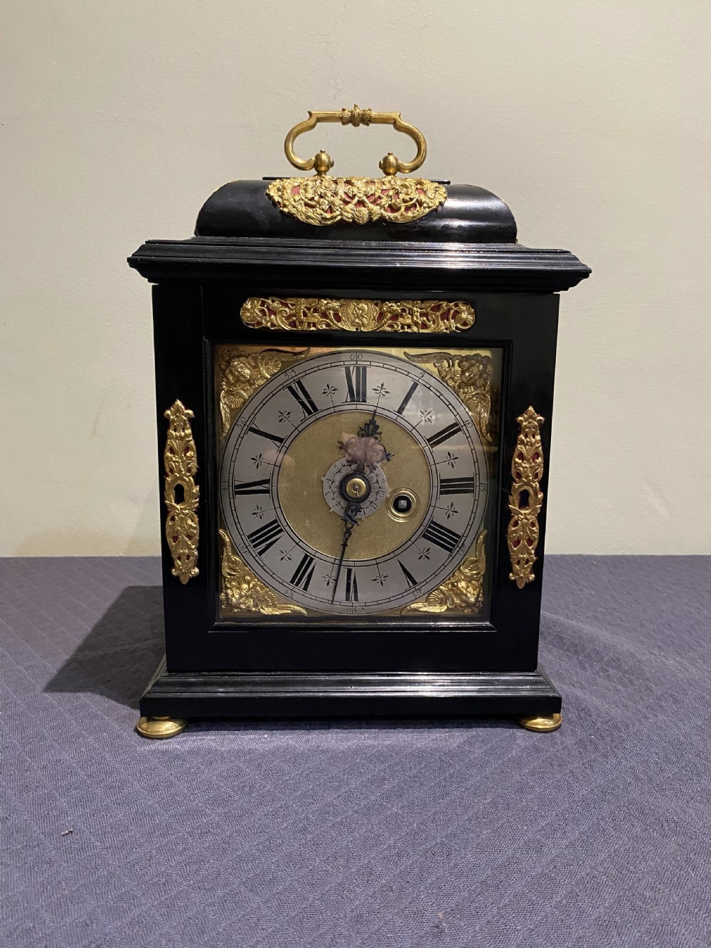 A late 17th-century black ebony bracket clock featuring decorative gilded brass mounts, a brass carrying handle, and a square glass face with a silvered chapter ring.