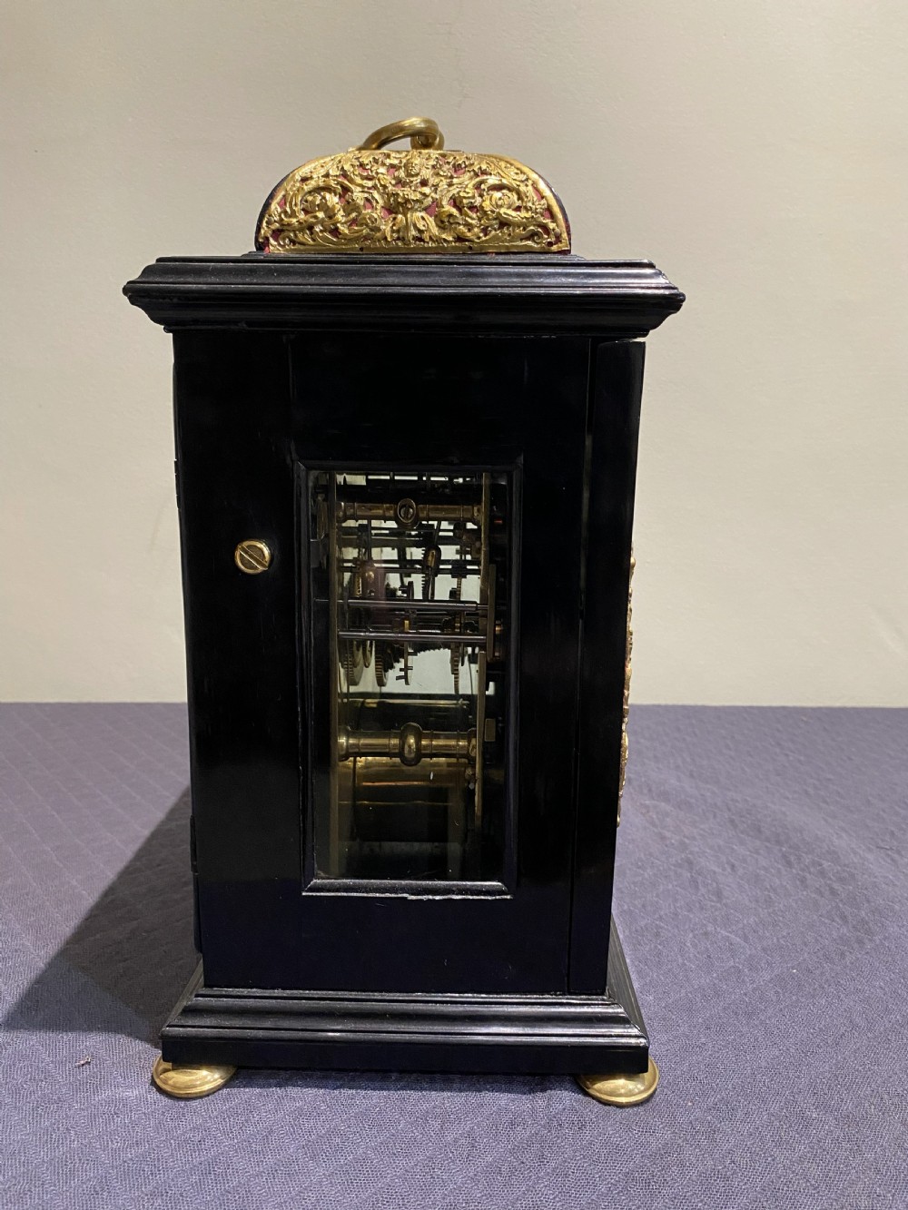 A late 17th-century black ebony bracket clock featuring decorative gilded brass mounts, a brass carrying handle, and a square glass face with a silvered chapter ring. View II