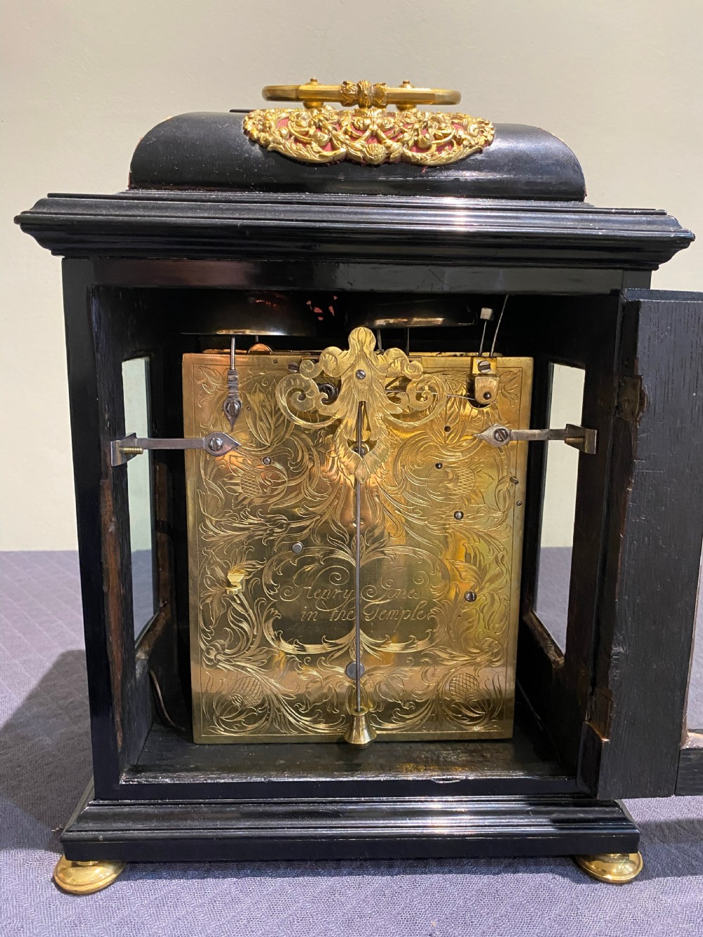 A late 17th-century black ebony bracket clock featuring decorative gilded brass mounts, a brass carrying handle, and a square glass face with a silvered chapter ring. View V