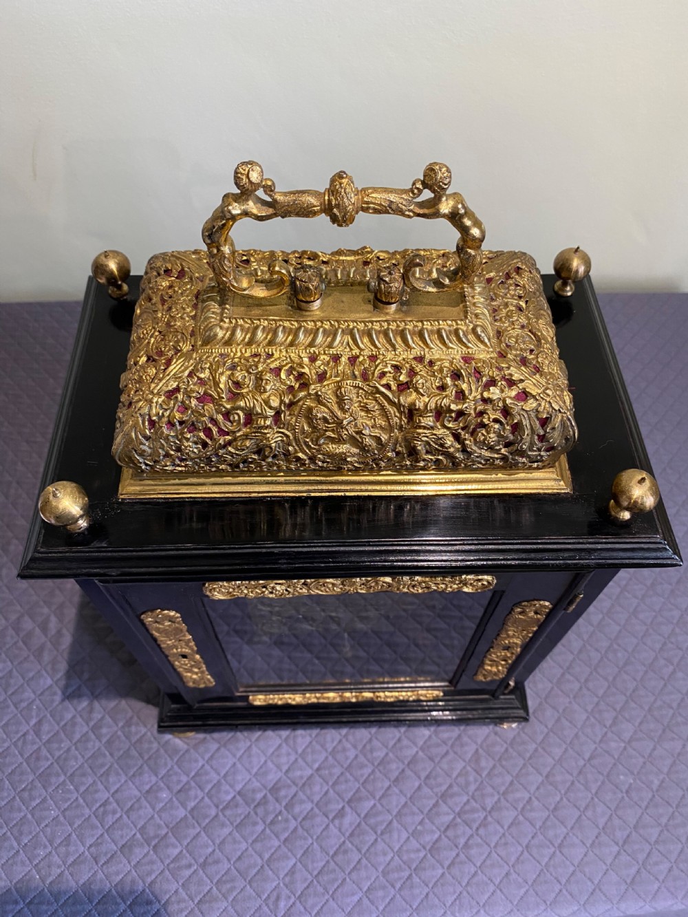 A late seventeenth-century William and Mary ebony table clock featuring an ornate gilded brass basket top with carrying handle and silvered chapter ring. View V