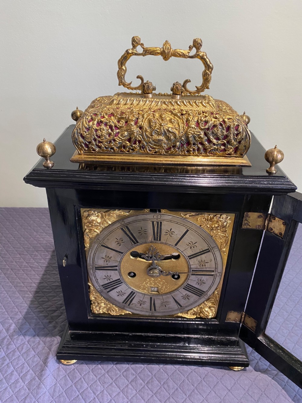 A late seventeenth-century William and Mary ebony table clock featuring an ornate gilded brass basket top with carrying handle and silvered chapter ring. View VI