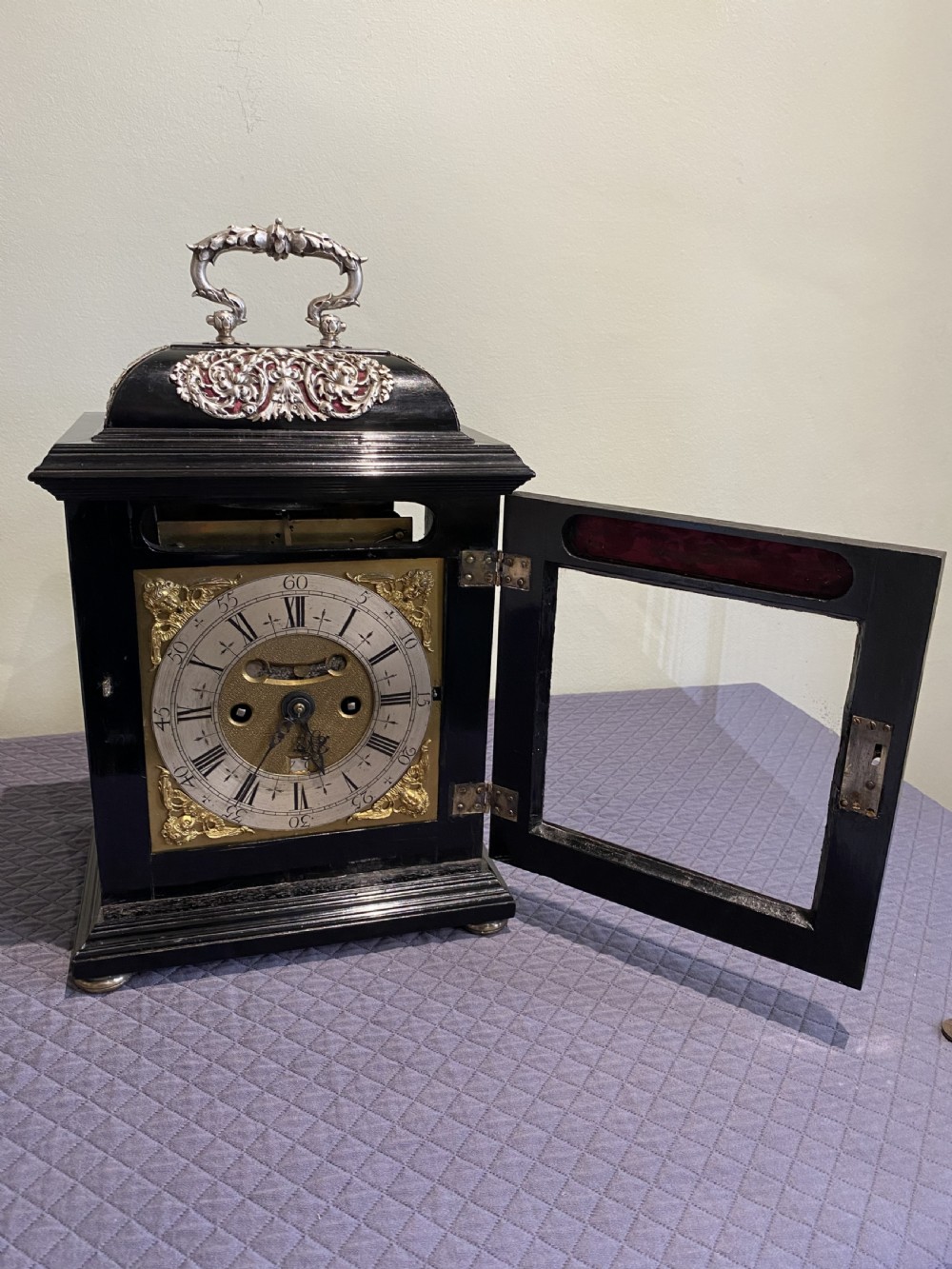 A late 17th-century black ebony table clock featuring repousse silver mounts, an ornate carrying handle, and a brass dial with silvered chapter ring. View II