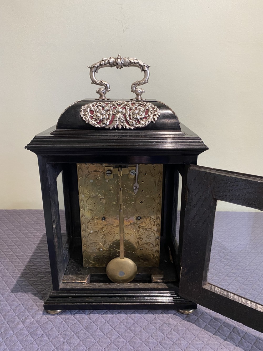 A late 17th-century black ebony table clock featuring repousse silver mounts, an ornate carrying handle, and a brass dial with silvered chapter ring. View V